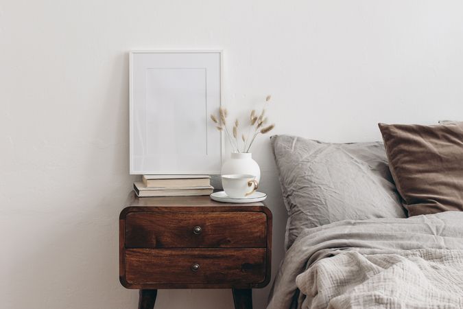 Vertical white picture frame mockup on old books. Elegant bedroom view. Brown, grey linen and velvet pillows, blanket. Wooden night stand with ceramic vase, dry grass and cup of coffee. Scandinavian interior.