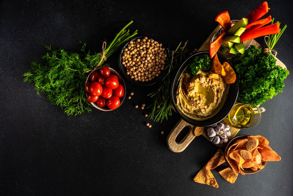 Top view of creamy hummus dip in bowl on board served with olive oil, veggies and chips
