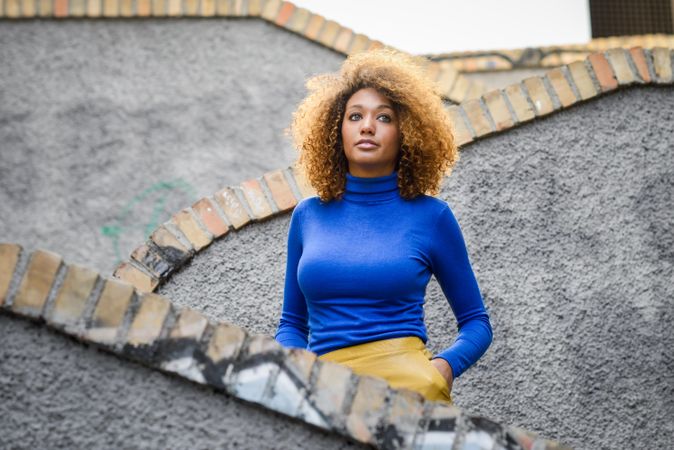 Nonchalant female with curly hair wearing bright blue shirt standing on outdoor stairs