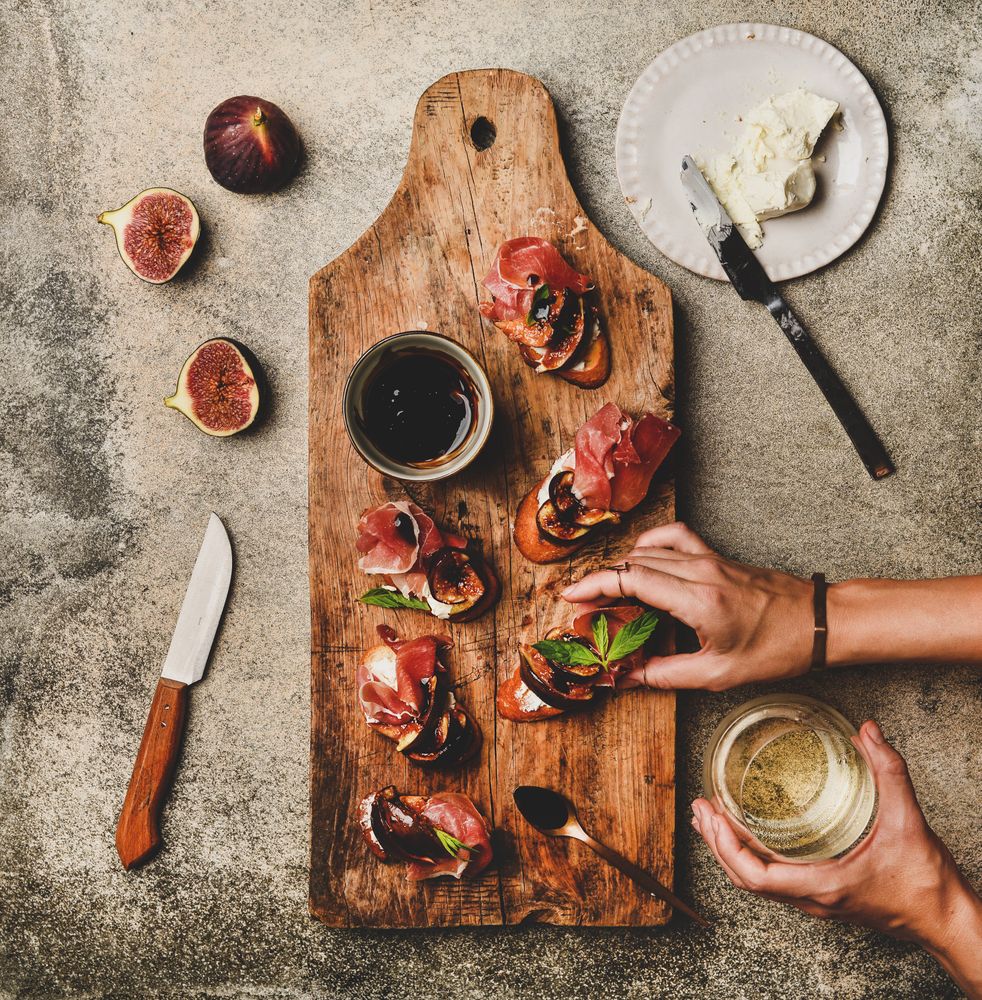 Hands with canapé of prosciutto, goat cheese and grilled figs, white wine, square crop