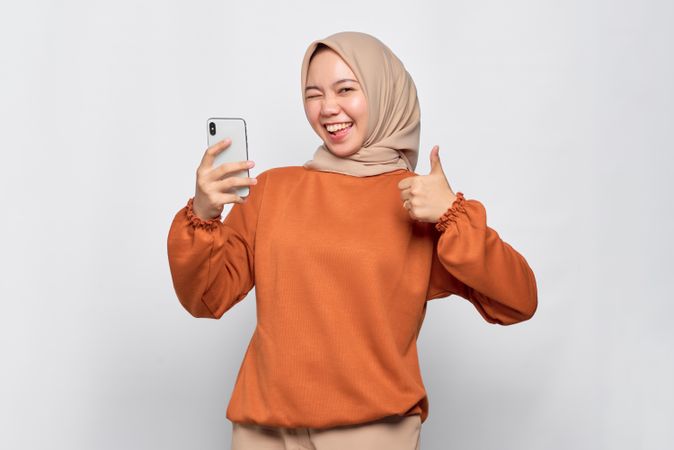 Happy Muslim woman in headscarf and orange shirt with phone and giving thumbs up