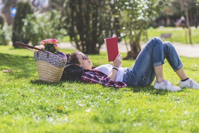 Side view of a hipster happy woman lying on grass in sunny day at park while reading a red book