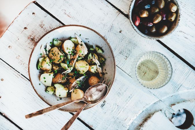 Greek style potato salad with serving utensils, olive bowl and wine, top view