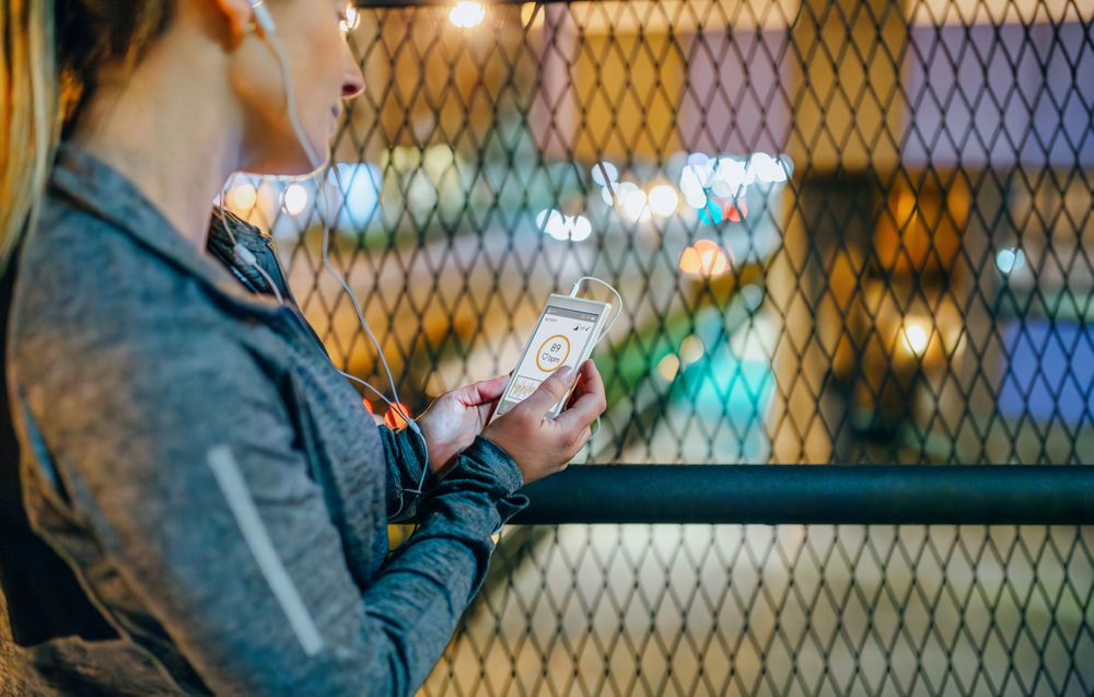Female runner looking screen of mobile phone with heart rate monitor after training on city at night