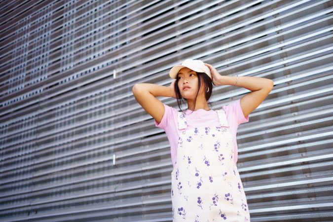 Chinese woman in floral coveralls looking away while holding her cap in front of metal wall