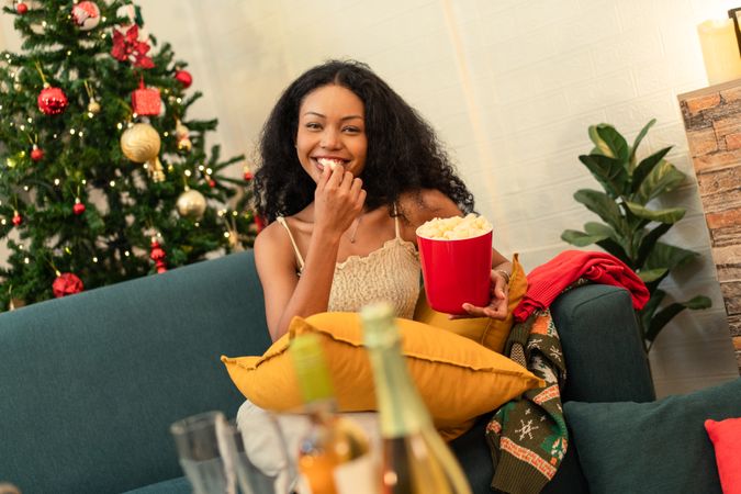 A Black woman eating popcorn while relaxing on a sofa on Christmas holiday