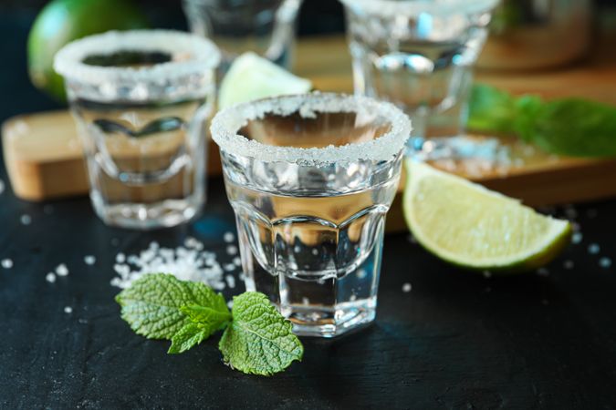 Tequila shots with salt, lime slices and mint on dark table