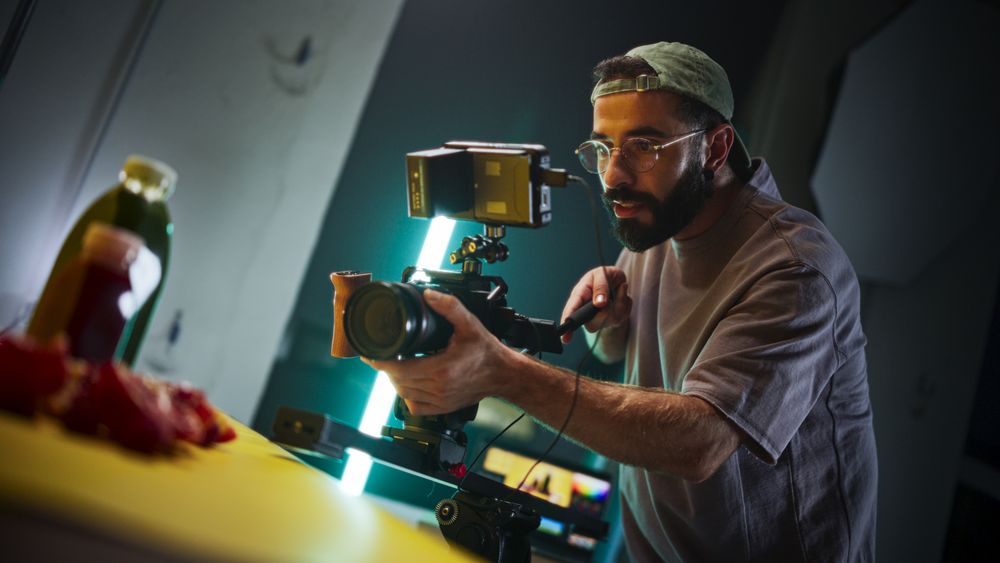 Focused Male Cinematographer With Beard And Glasses Adjusts Professional Camera On Film Set, Capturing Vibrant, Dynamic Footage For A Documentary Or Advertising Project Under Studio Lighting.