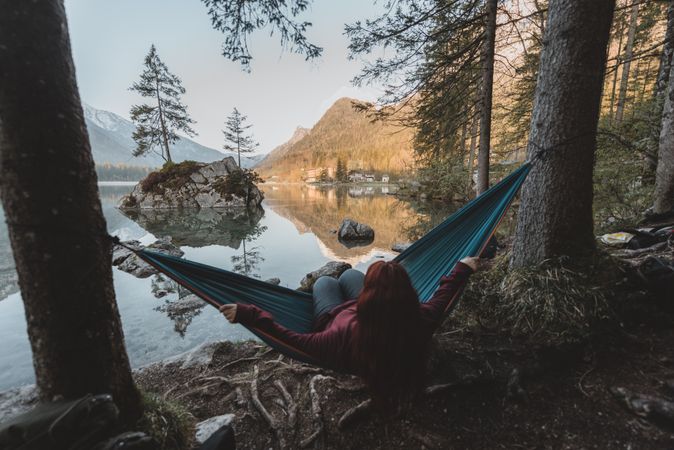 Woman in red jacket sitting on blue hammock near lake during daytime
