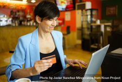 Smiling female in trendy blue jacket sitting in cafe with laptop and credit card 56G2AV