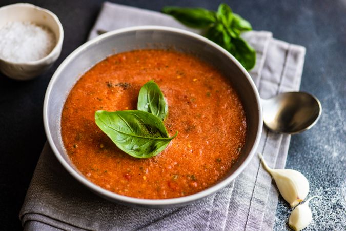 Grey bowl of gazpacho soup with basil leaves on napkin