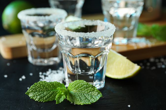 Tequila shots with salt, lime slices and mint on dark table