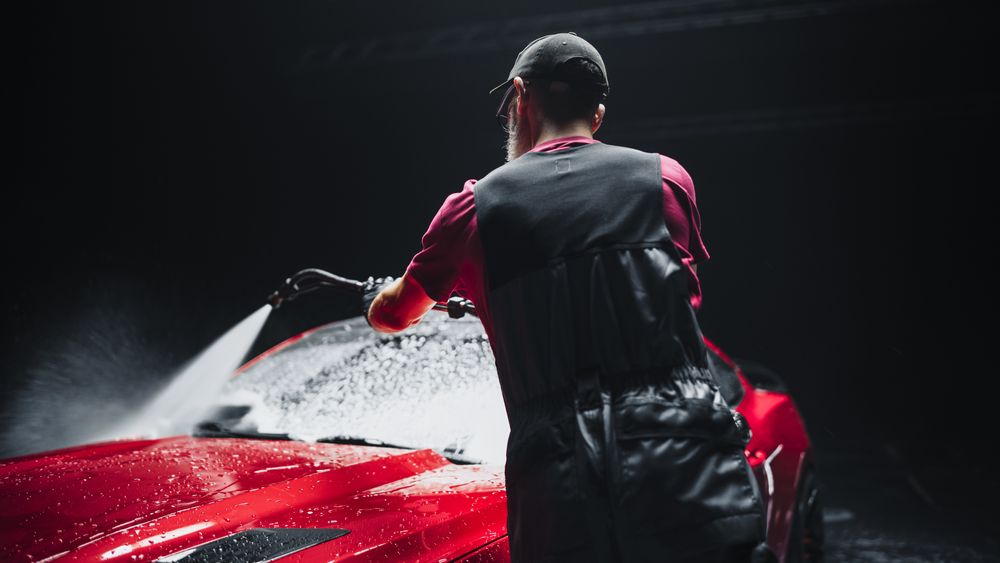 Automotive Detailer Washing Away Shampoo from the Windshield with a Water High Pressure Washer.