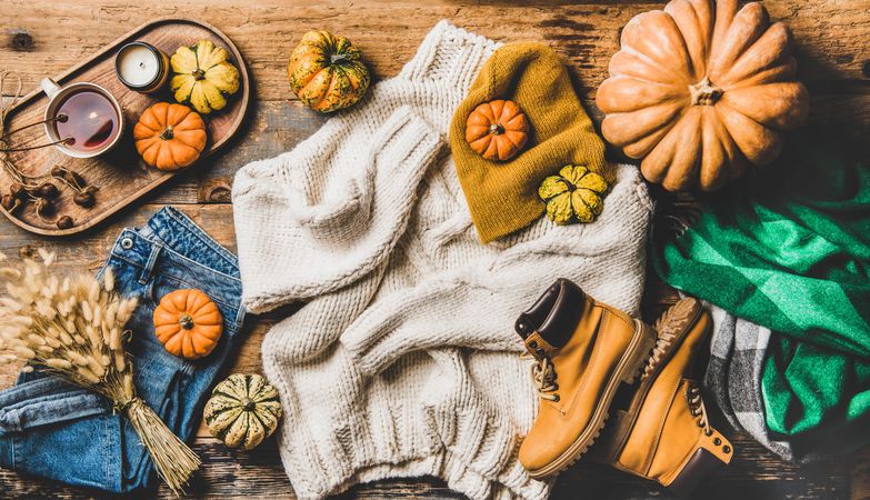 Flat-lay of beige sweater, blue jeans, green scarf, hat, yellow boots, pumpkins, candle and tea