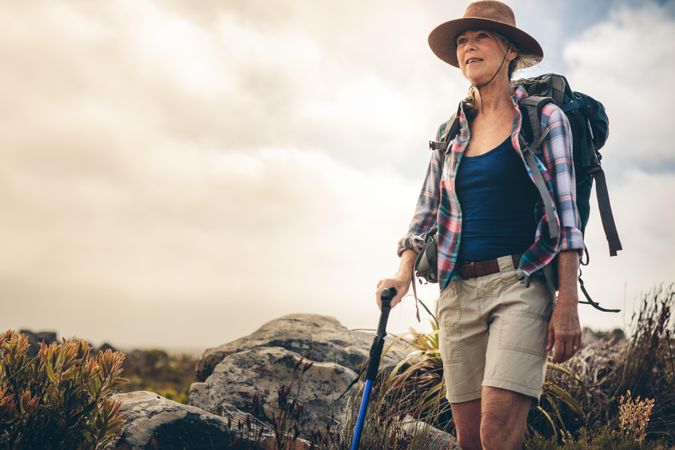 Older woman wearing hat and backpack trekking in the countryside holding a hiking pole
