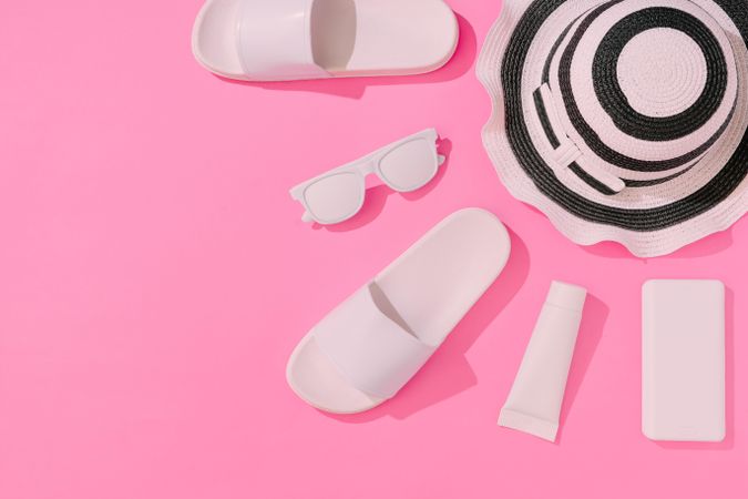 Summer objects of flip flops, sunglasses, smartphone, sunscreen lotion, hat on pink background