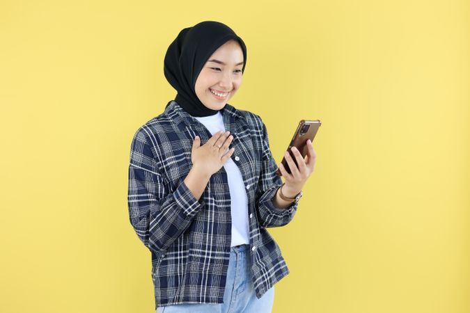 Happy Excited Asian Muslim Woman Wearing Hijab Holding Smartphone while TOuch Her Chest Smiling Cheerfully, Isolated on Yellow Background.