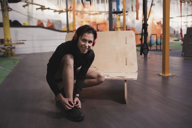 Smiling woman with headphones lacing shoe in gym