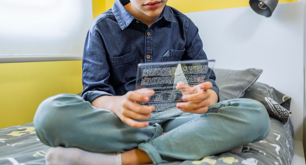 Unrecognizable boy using futuristic transparent tablet to read ebook sitting over his bed