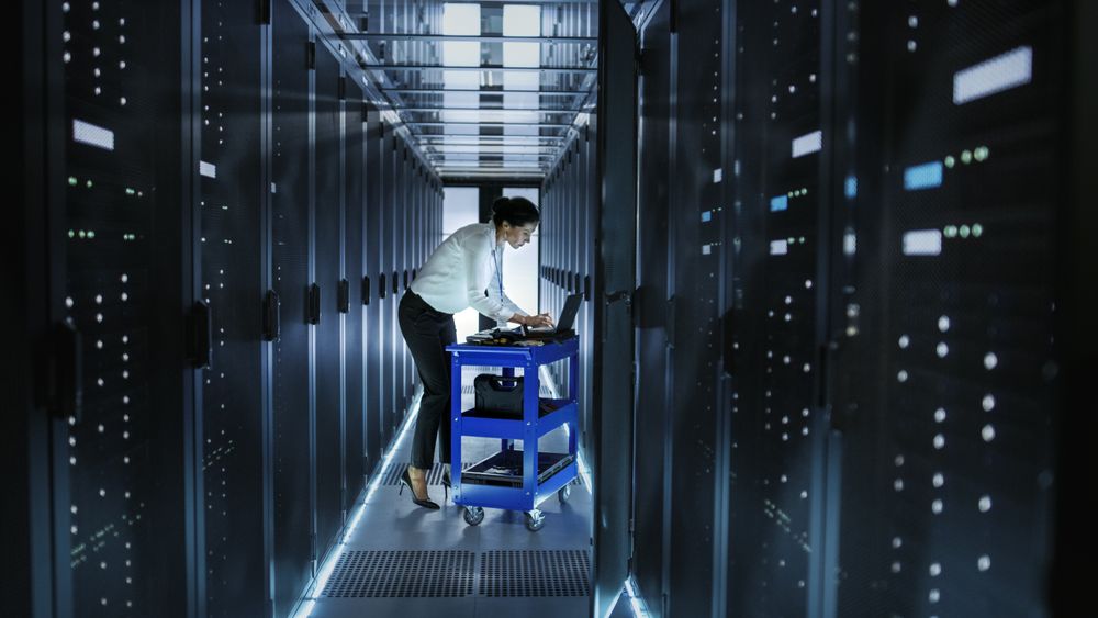 Female IT Engineer Works on a Laptop and Scans and Swaps Hard Drives That are Lying on a Cart.