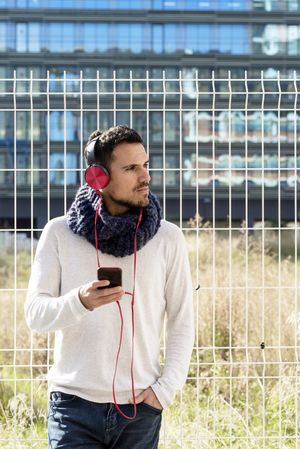 Man walking in scarf leaning back on metallic fence and looking up from using smartphone