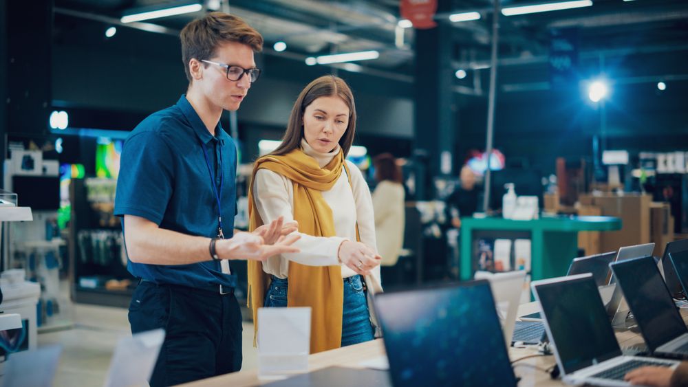 Young Woman Having a Conversation with a Salesperson in Home Electronics Store while Selecting a Laptop.