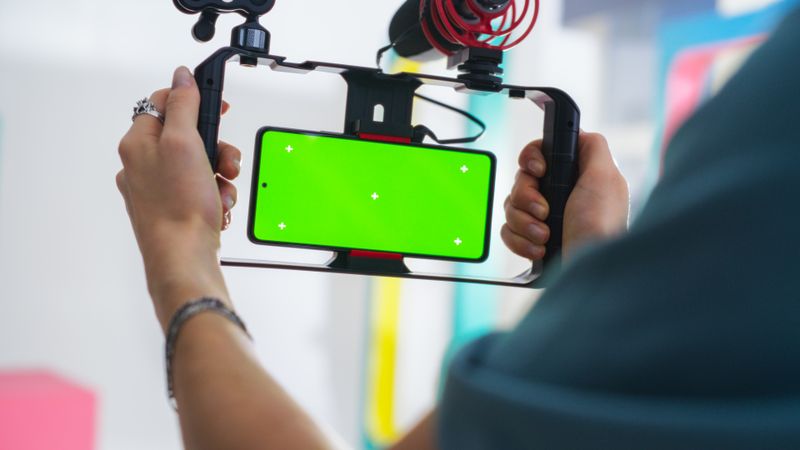 Close-up Of Young Male Cinematographer Using Smartphone With Green Screen Mounted On Gimbal For Video Production In Vibrant, Modern Studio Setting.