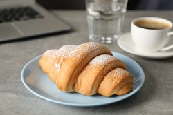 Blue plate with croissant, coffee and water next to computer on grey table, close up