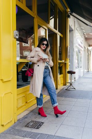 Young cheerful female leaning on house with smartphone