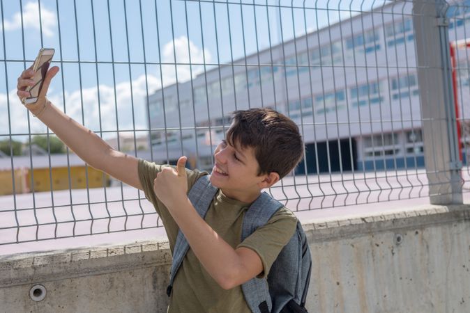 Smiling boy giving thumbs up while taking selfie on smartphone outside of school yard