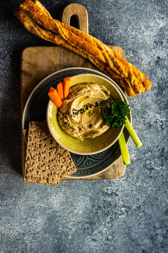 Top view of creamy hummus with bread sticks and rye crackers on grey kitchen counter