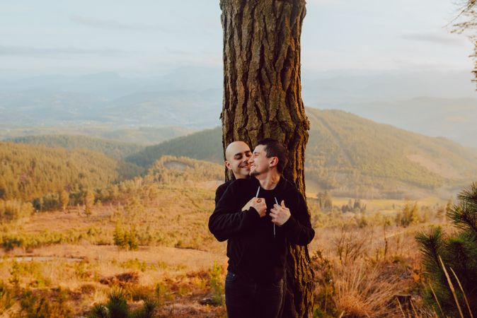 Smiling men hugging near tree in forest on hill
