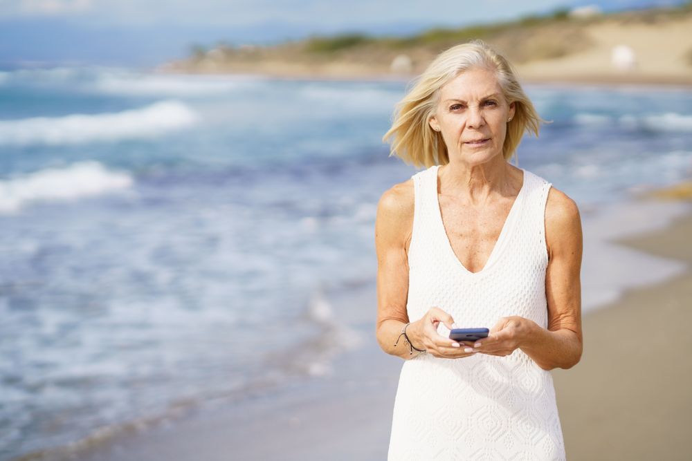 Serious older woman using her phone on a rocky beach