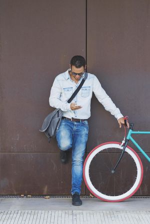 Male in sunglasses standing with red and green bicycle in front of wall and using phone, vertical
