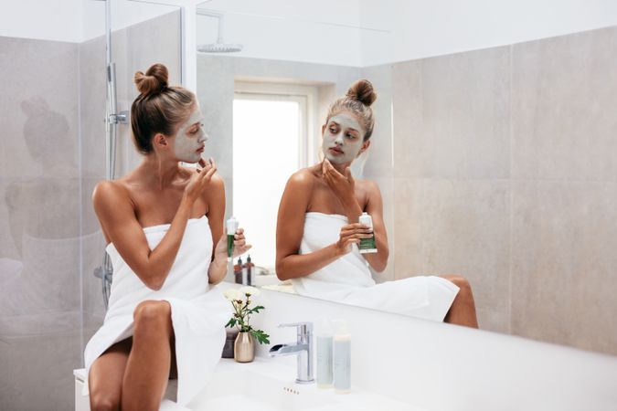 Young woman applying facial mud clay mask to her face in bathroom.