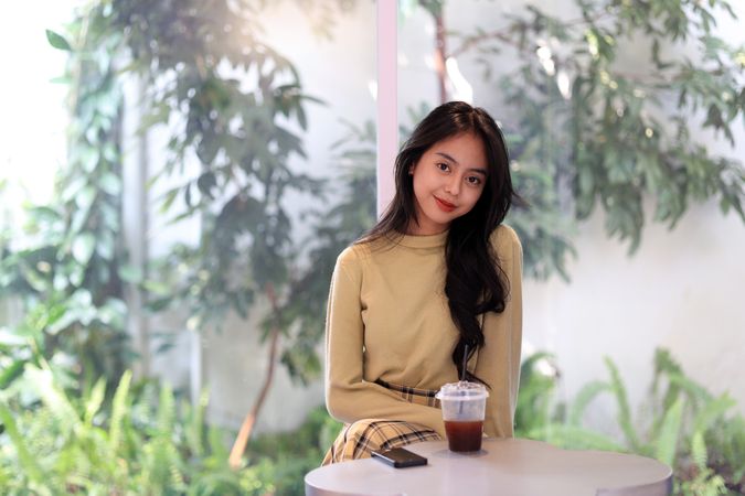Young Asian Woman Smiling Behind a cup of Ice Coffee Americano on a White Table.
