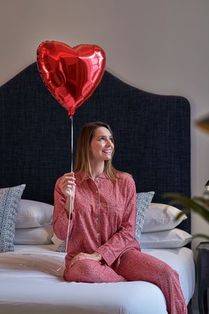 Cheerful attractive woman sitting on bed with red heart balloon
