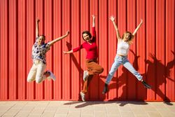 Three women jumping for joy in front of red wall on sunny day