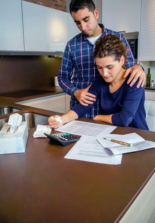Couple supporting each other as they go through their bills together in the kitchen, vertical