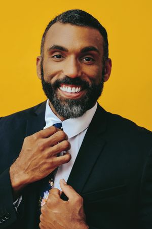Smiling Black male in suit adjusting his floral tie in yellow studio