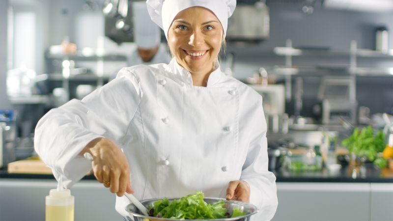 In a Famous Restaurant Female Cook Prepares Salad and smiling on a camera. She Works in a Big Modern Kitchen.