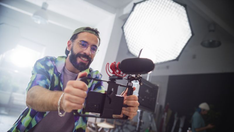 Young Male Videographer With Beard, Wearing Colorful Shirt And Cap, Enthusiastically Operates Camera In Studio Setting, Capturing Content For Advertising Or Movie Production.