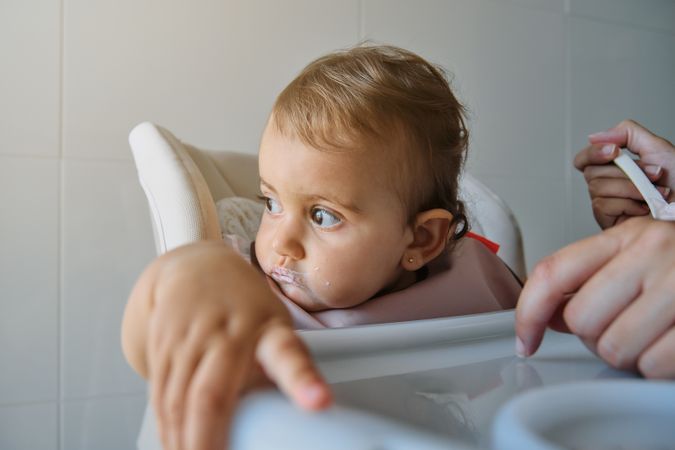 Unrecognizable woman feeding daughter on high chair with spoon