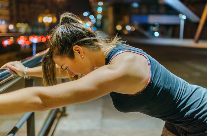 Portrait of female young athlete stretching using banister after training at night in city