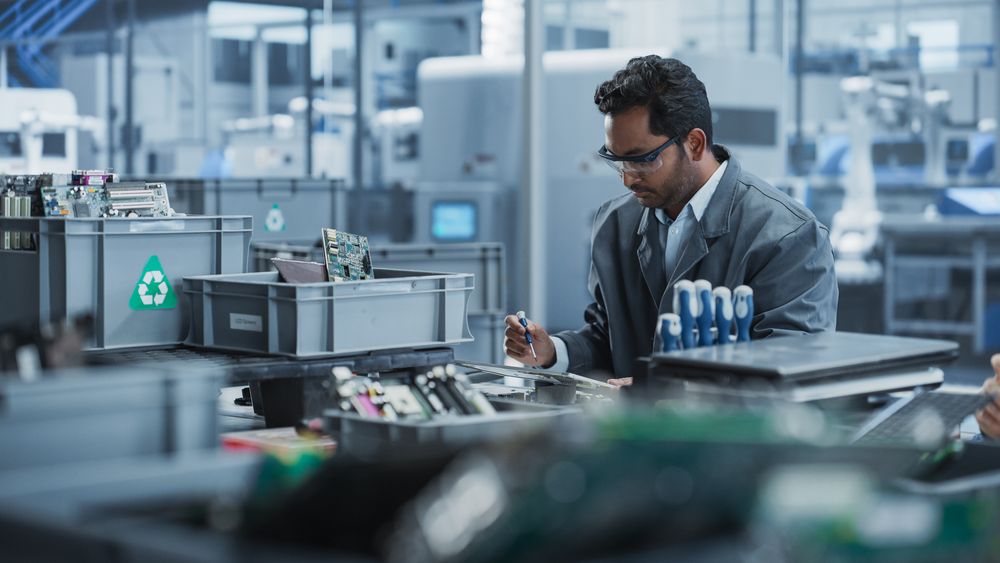 Indian Male Worker Disassembling Laptops To Recycle Electronic Components For Microchip Production At An Electronics Factory With Robotic Arms.