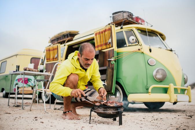 Tanned man cooking on camping stove beside parked mini van