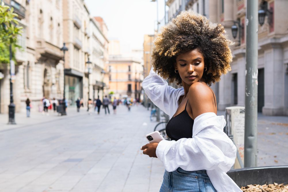 Black woman touching hair while standing on street with smartphone