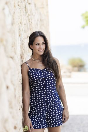 Latina woman in blue dotted dress leaning on wall outside and looking away, vertical