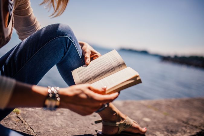 Cropped image of woman in blue denim jeans reading book by shoreline