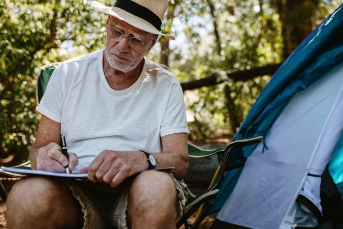 Older man sitting in front of a tent and writing in a book
